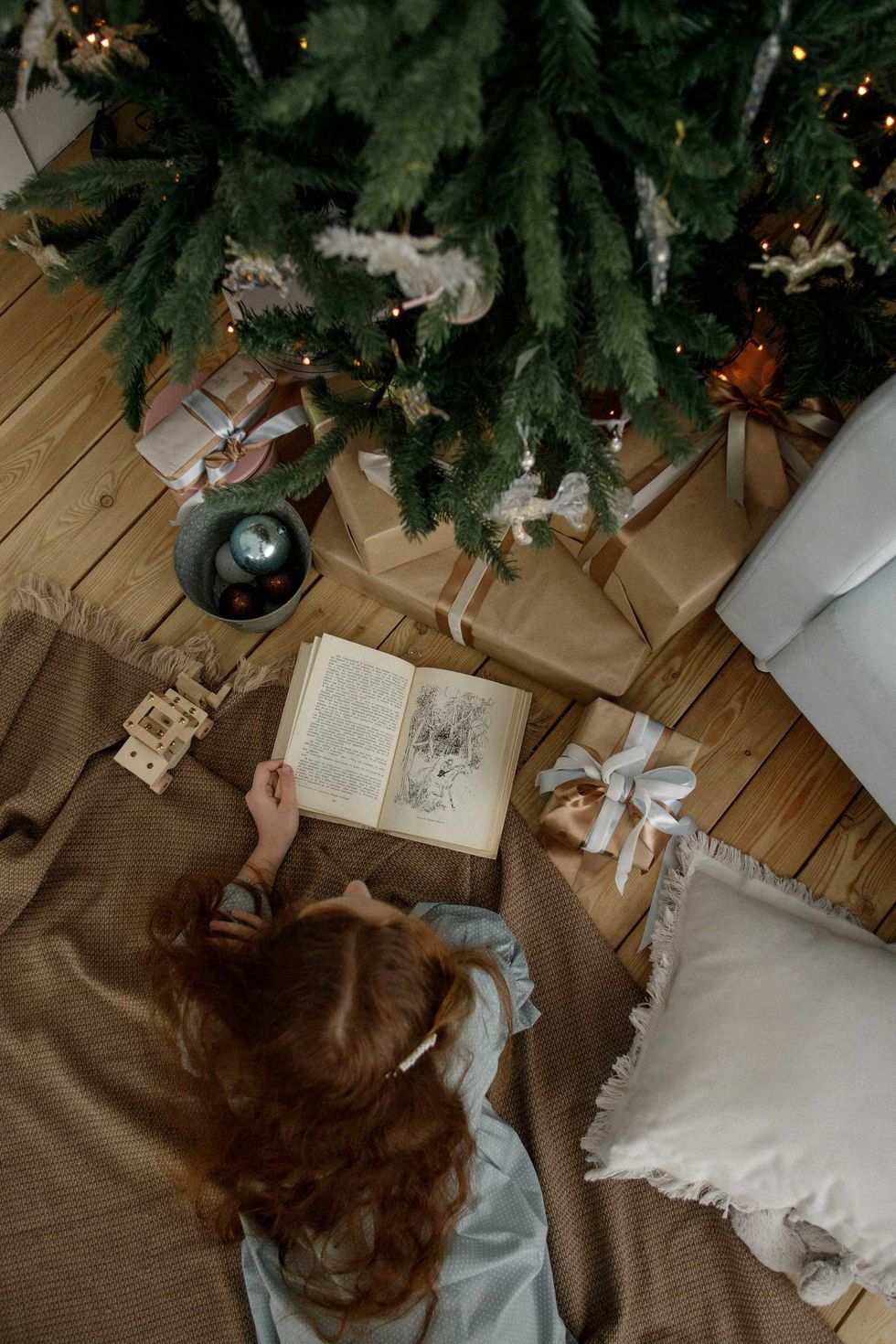 Person reading under a decorated Christmas tree with gifts on a wooden floor.