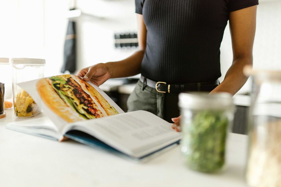 Person reads a cookbook in kitchen with jars of ingredients nearby.