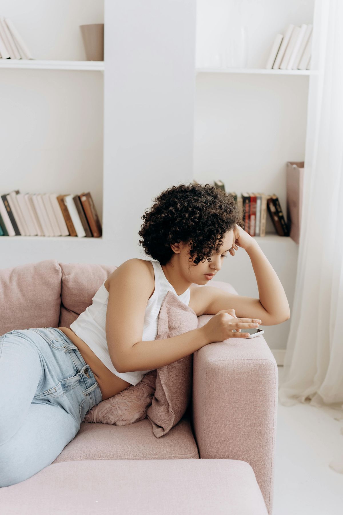 Person reclining on a pink sofa using a smartphone in a cozy, book-filled room.