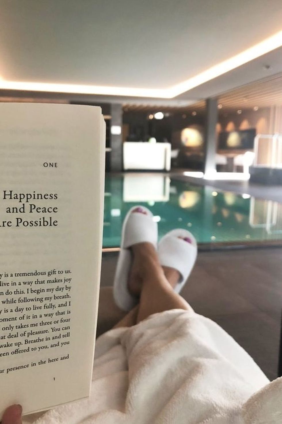 Person relaxing by a pool with a book in hand, wearing slippers and a robe.