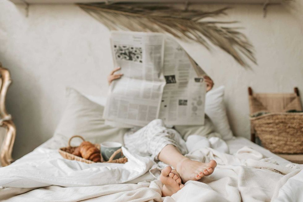 Person relaxing in bed with a newspaper, croissants, and coffee on a tray.