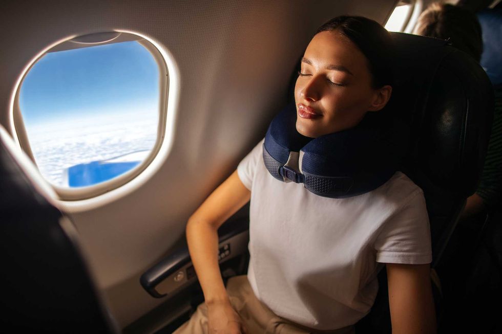 Person relaxing on a plane with a neck pillow, looking out the window at clouds.