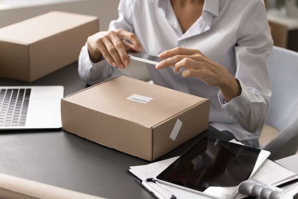 Person sealing a cardboard box at a desk with a laptop and tablet.