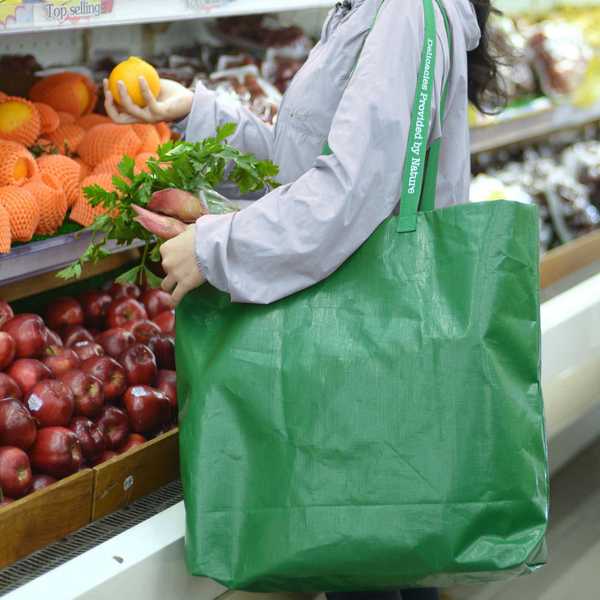Person shopping with a large green tote bag, holding fruits and vegetables.