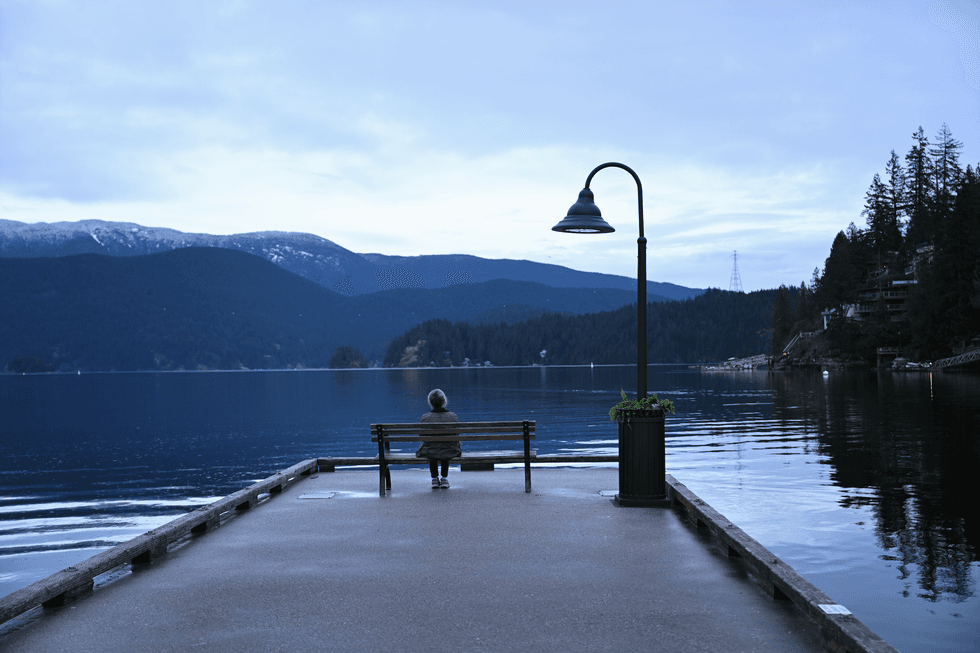 Person sits on a bench at a pier, overlooking a calm lake and mountains at dusk.