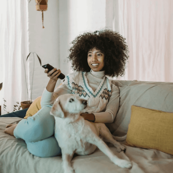 Person sitting on a couch with a dog, holding a remote, smiling at the TV.