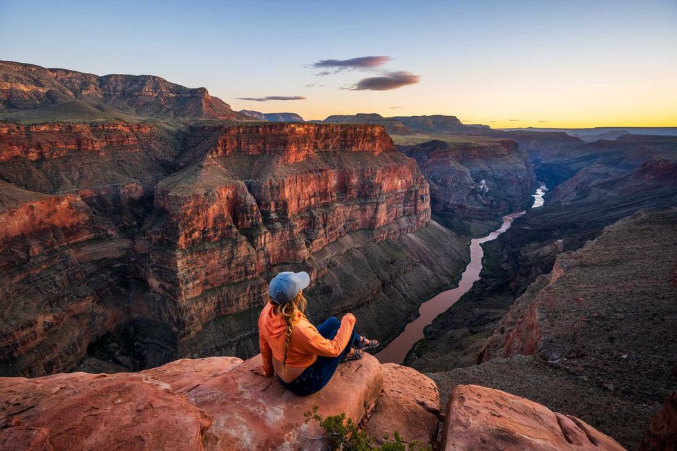 Person sitting on cliff edge, overlooking a vast canyon at sunset.
