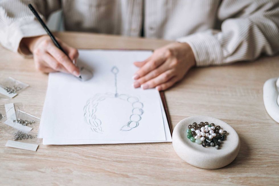 Person sketching jewelry design with beads on a table.