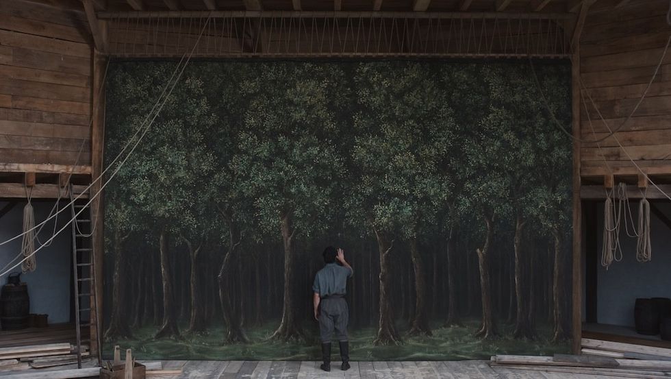 Person standing in front of a painted forest backdrop in a wooden theater setting.