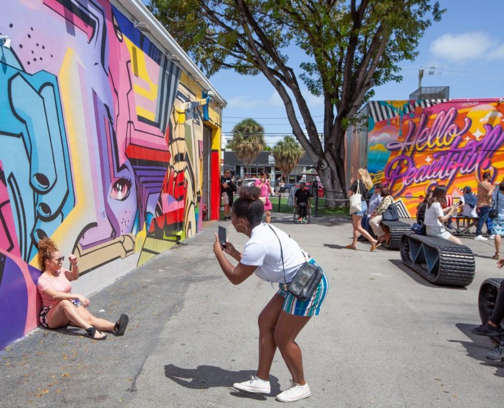 Person taking a photo of a colorful street mural and another person posing in front of it.