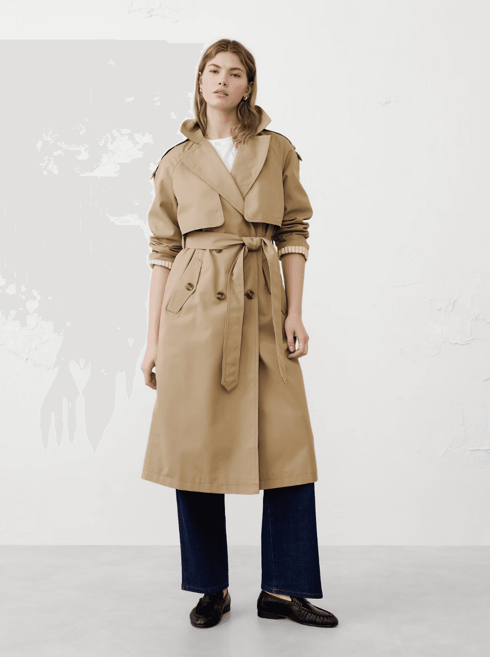 Person wearing a beige trench coat, white top, jeans, and black loafers.