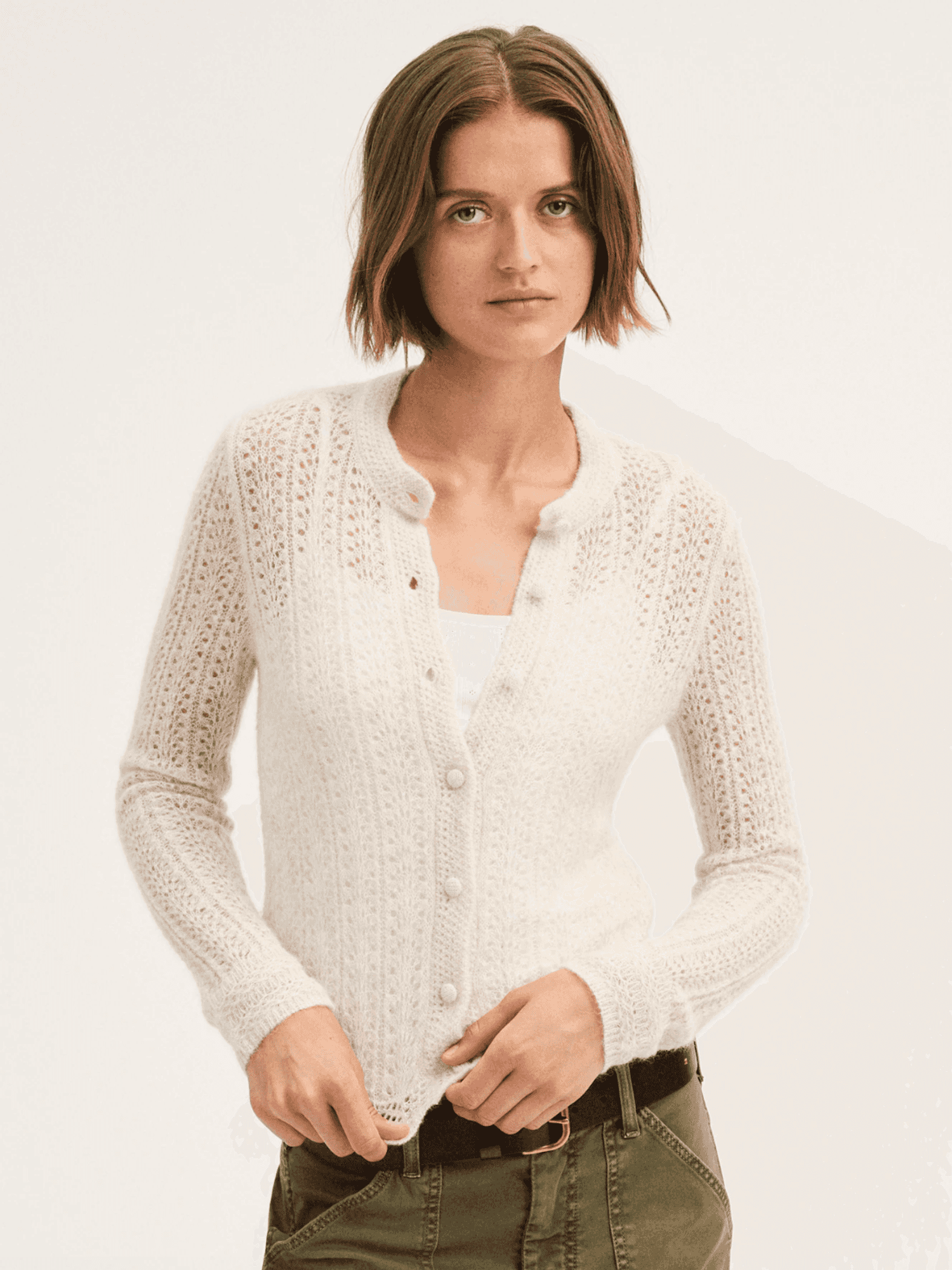 Person wearing a white knit cardigan and green pants, standing against a plain background.