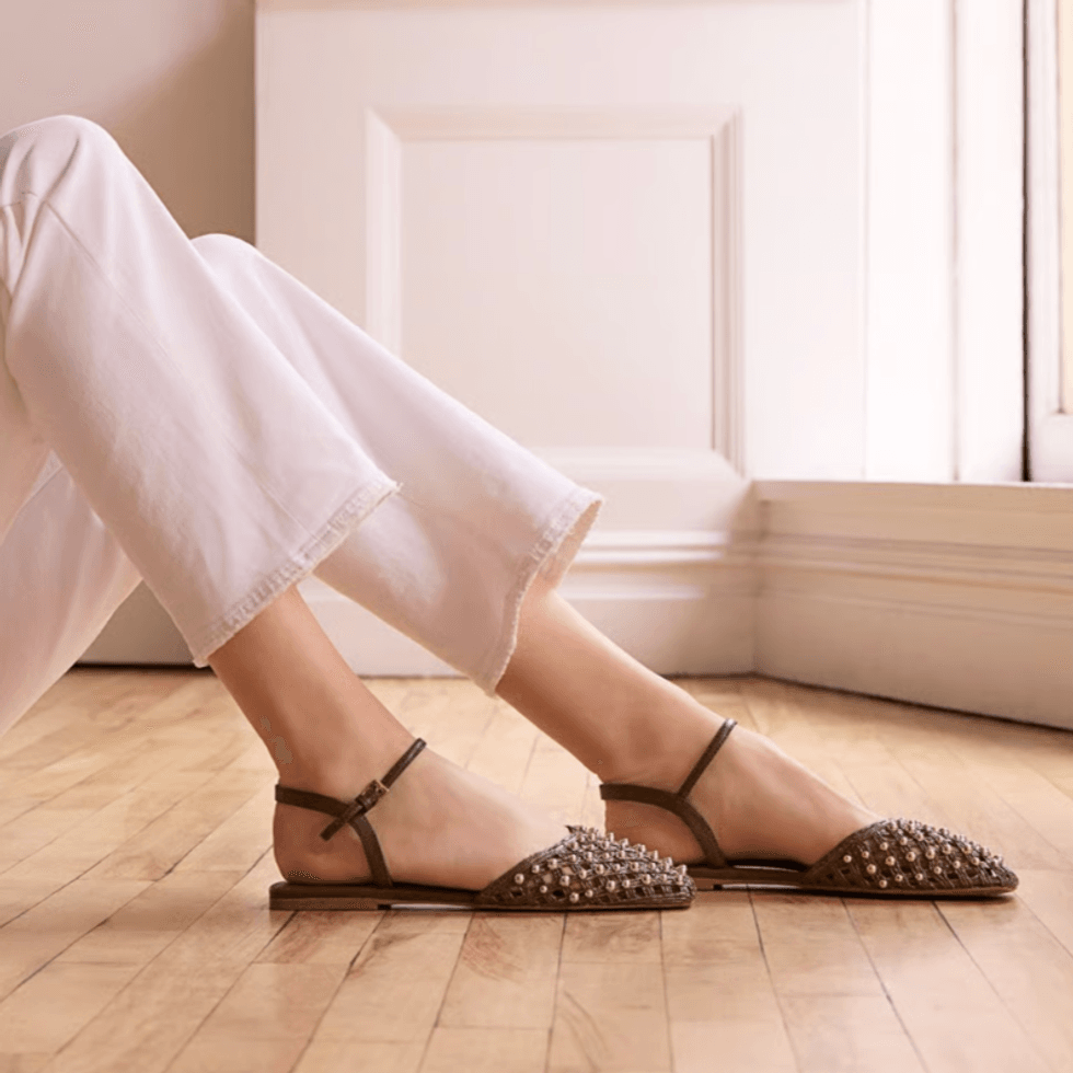Person wearing studded flats and white pants sitting on a wooden floor.