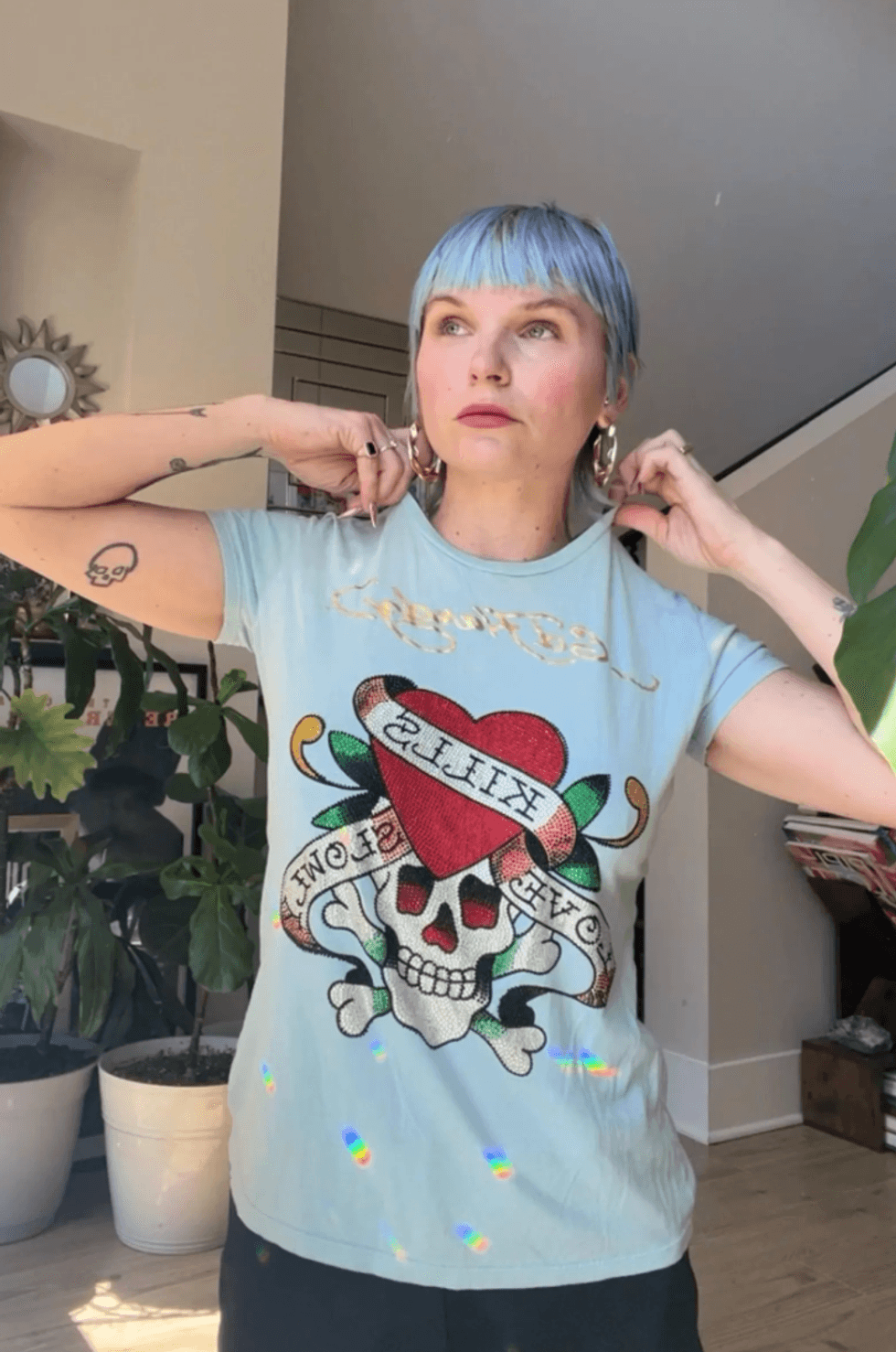 Person with blue hair in a graphic t-shirt, adjusting earrings at home.