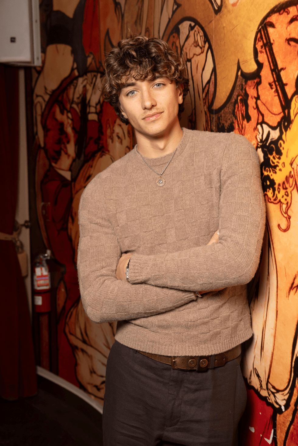 Person with curly hair leans against a colorful mural, wearing a beige sweater and necklace.
