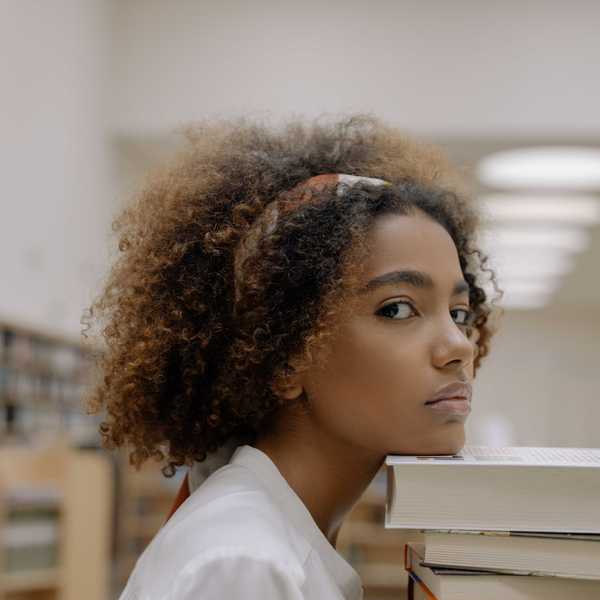 Person with curly hair leans on stacked books in a library setting.