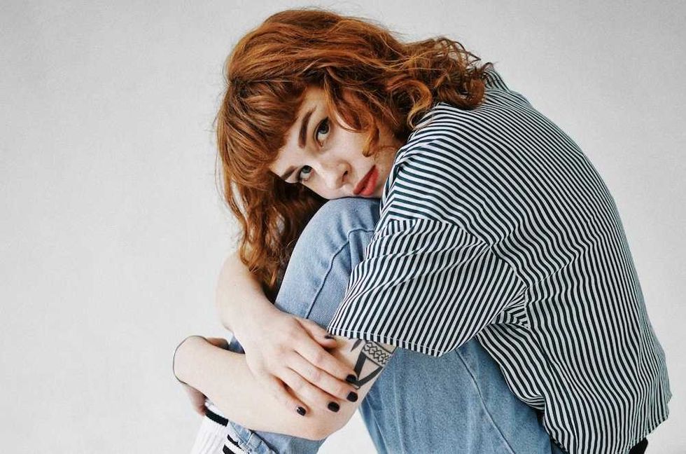 Person with curly red hair in striped shirt, hugging knees while sitting on floor.