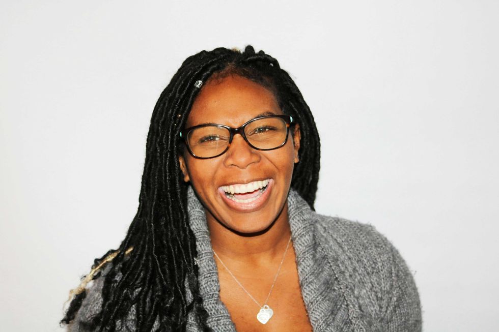 Person with glasses and long hair laughing, wearing a gray sweater and necklace.