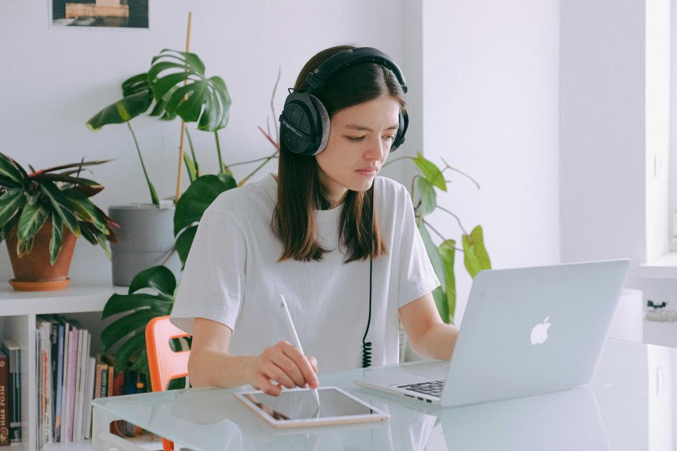 Person with headphones working on laptop and tablet in a bright room with plants.