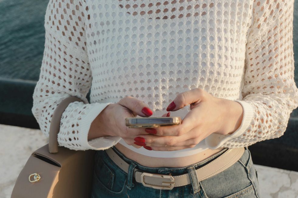 Person with red nails using a phone, wearing a white crochet top and jeans.