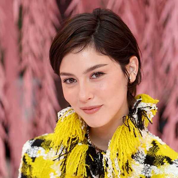 Person with short hair in a yellow and black patterned outfit against a pink feather backdrop.