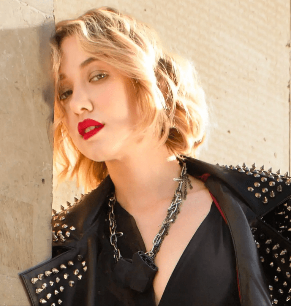 Person with wavy hair, red lips, wearing spiked leather jacket and necklace, leaning on wall.