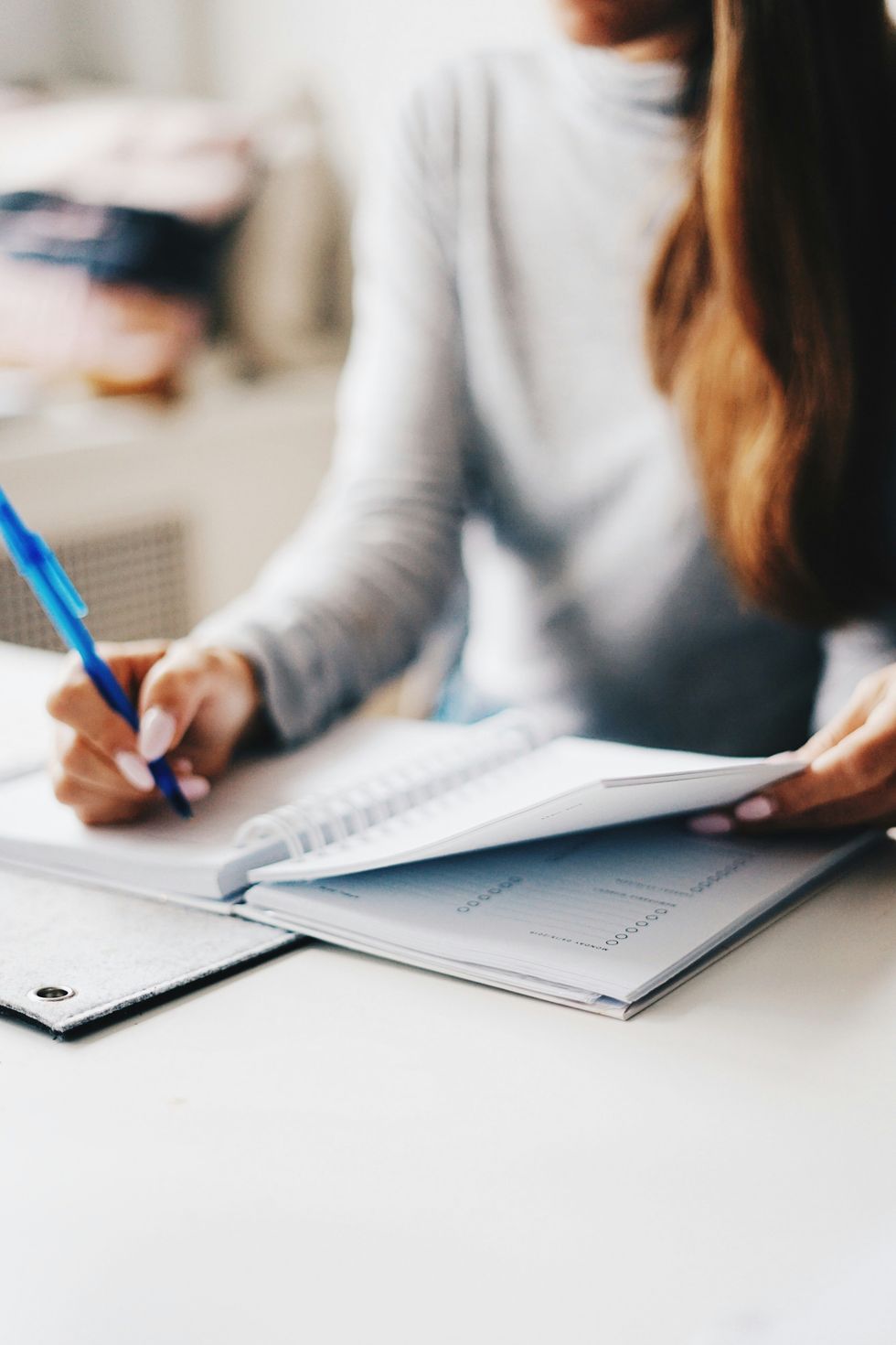 Person writing in a notebook with a blue pen on a white desk.