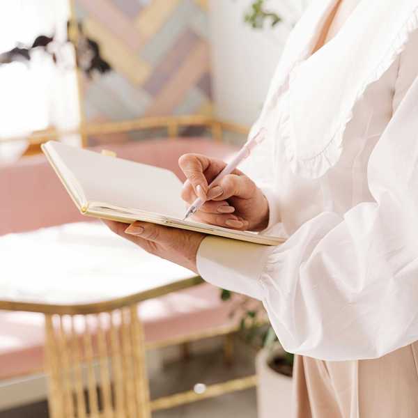 Person writing in a notebook with a pen, wearing a white blouse in a cozy room.