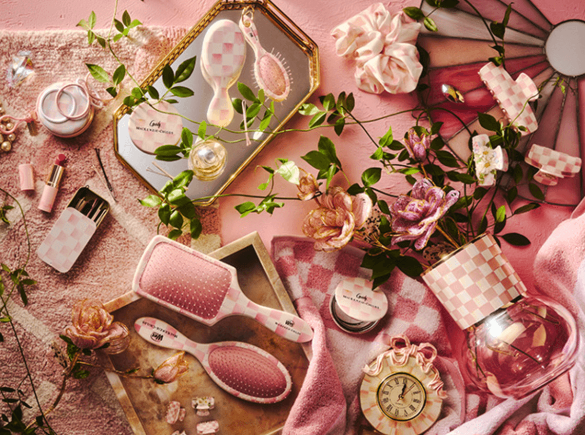 Pink beauty and decor items, including brushes, flowers, clock, and cosmetics on a soft pink surface.