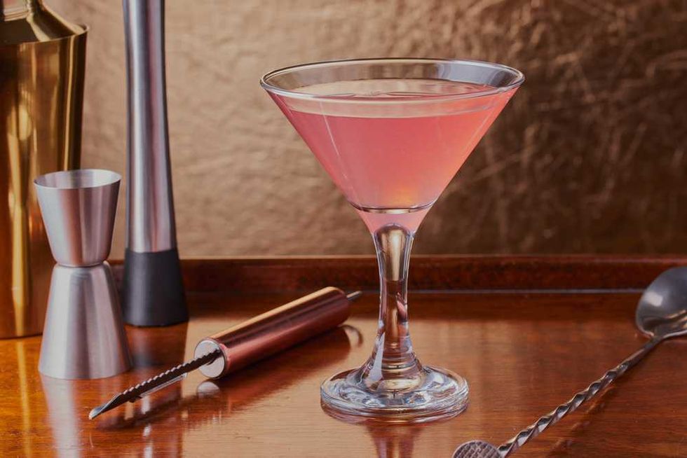 Pink cocktail in a martini glass with bar tools on a wooden tray.