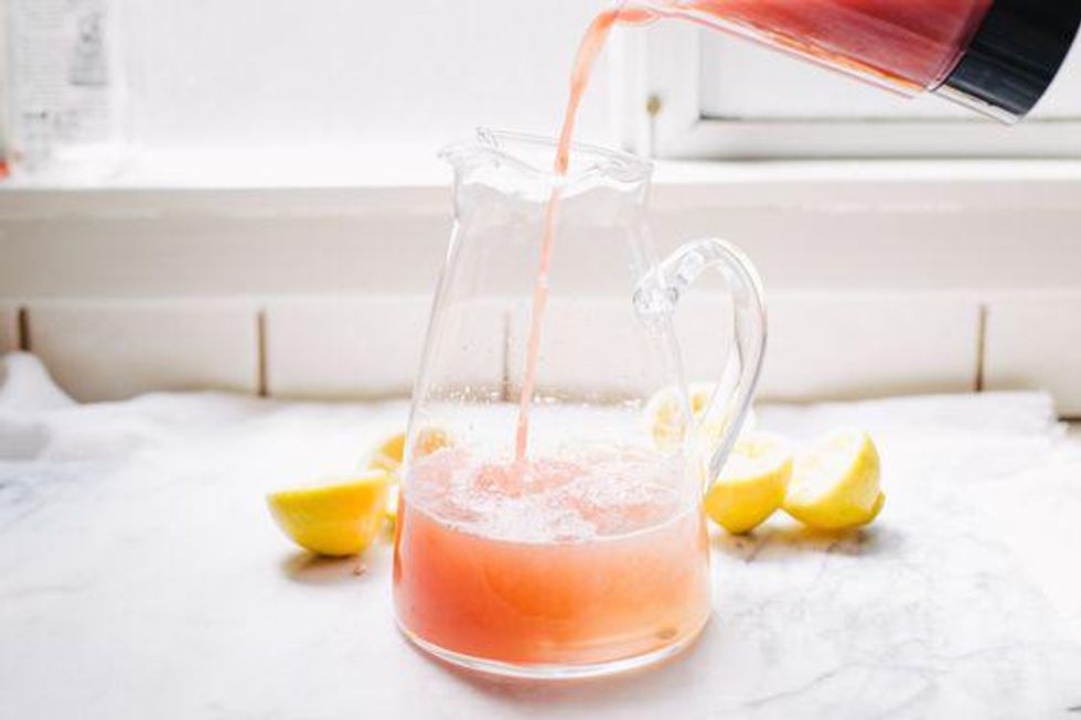 Pink lemonade being poured into a glass pitcher, with lemons nearby.