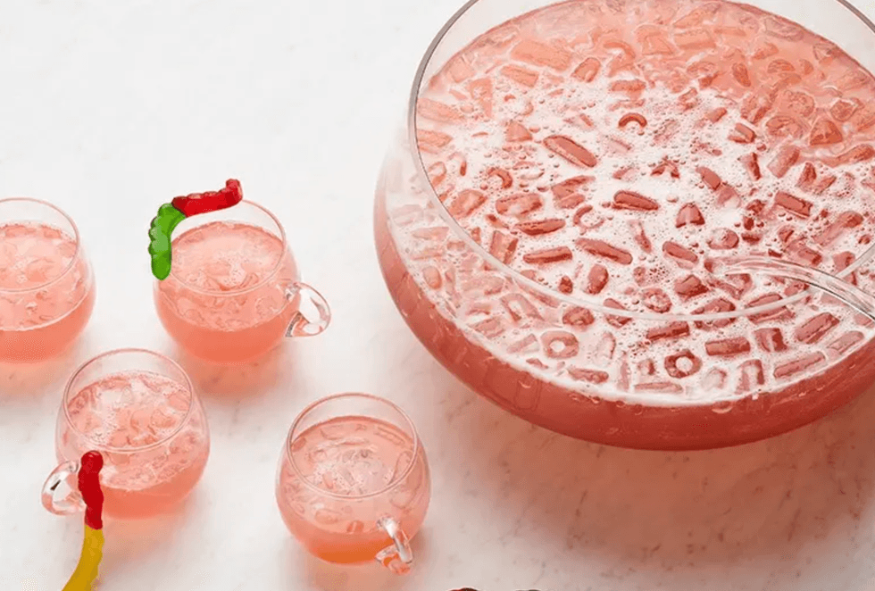 Pink punch bowl with cups, gummy worms as garnish, on a white surface.