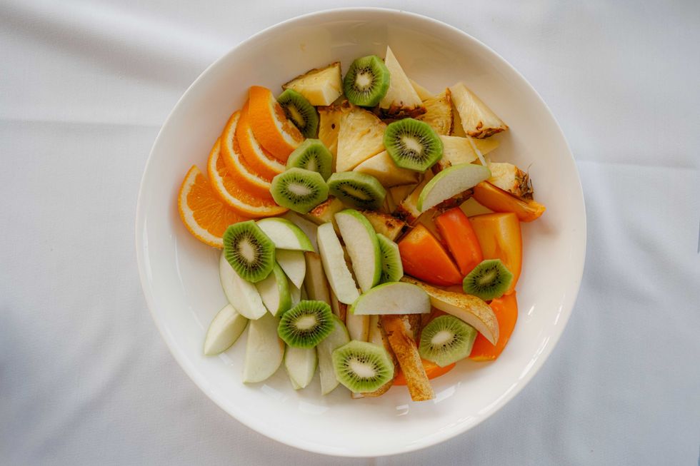 Plate of sliced fruits: orange, kiwi, apple, pineapple, and persimmon on white background.