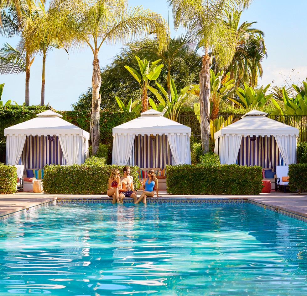 Poolside scene with three people and cabanas under palm trees.