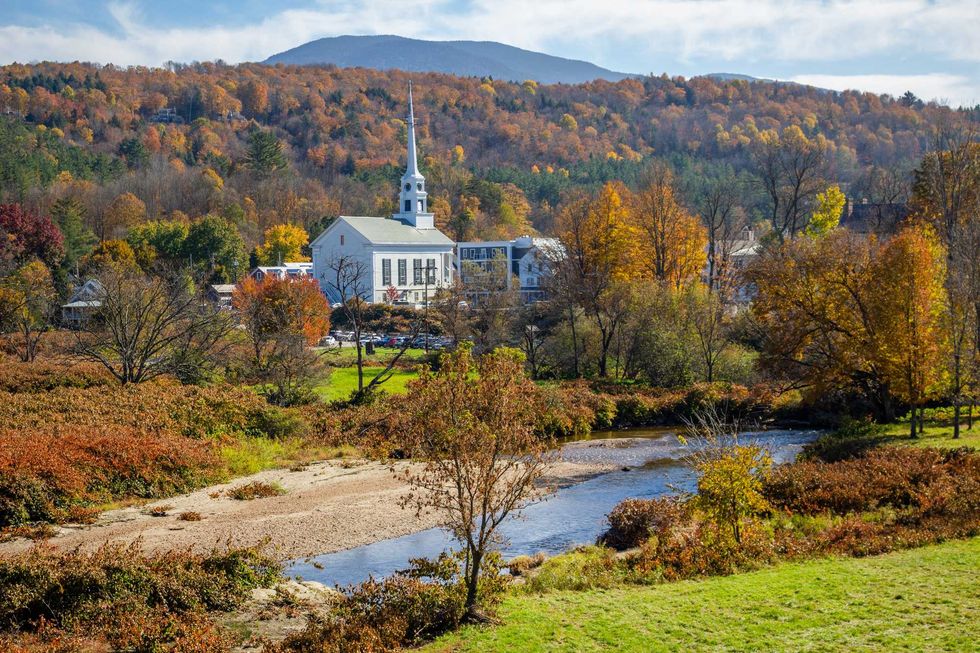Quaint church steeple amidst colorful autumn trees and a gentle stream at the base of a hill.