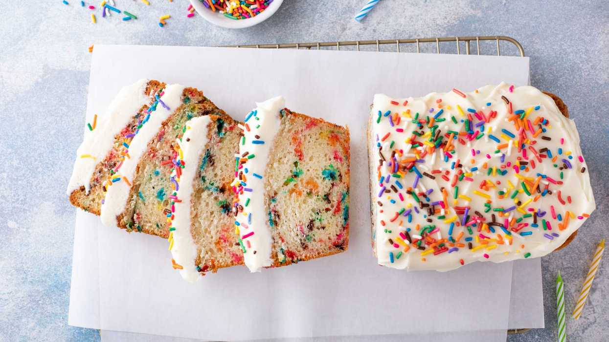Rainbow sprinkle loaf cake with white frosting, partially sliced, on parchment paper.