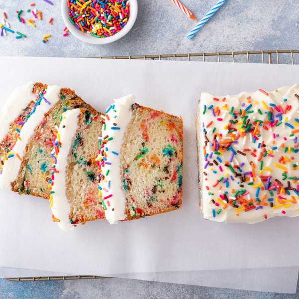 Rainbow sprinkle loaf cake with white frosting, partially sliced, on parchment paper.