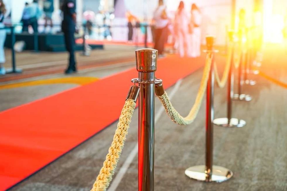 Red carpet event with stanchions and ropes, bright sunlight in the background.