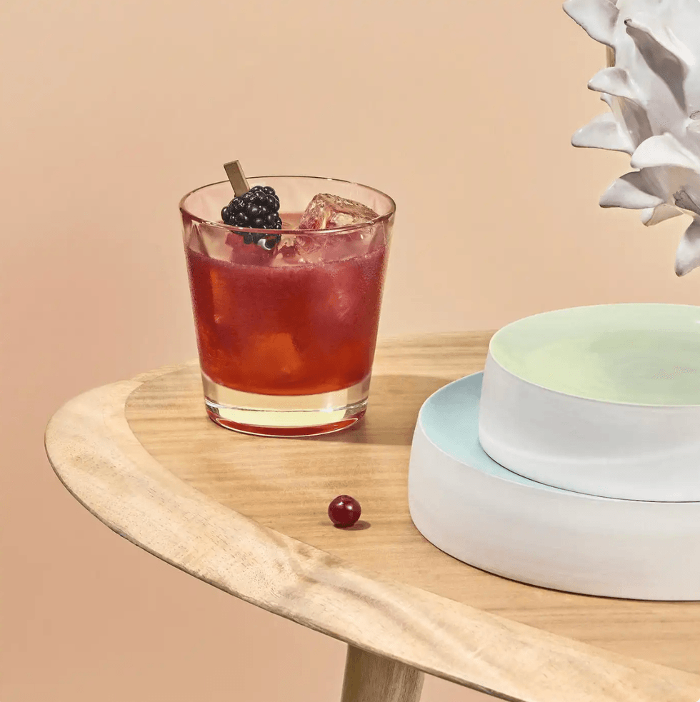 Red cocktail with blackberry on wooden table next to plates and sculpture.