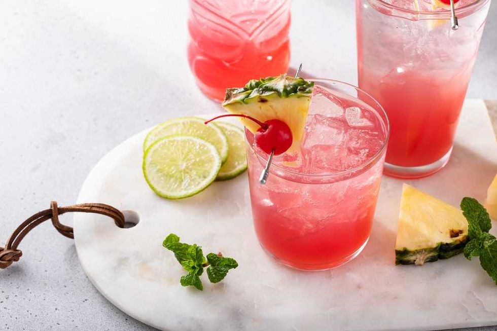Red cocktail with pineapple, cherry garnish, lime slices, and mint on a marble board.