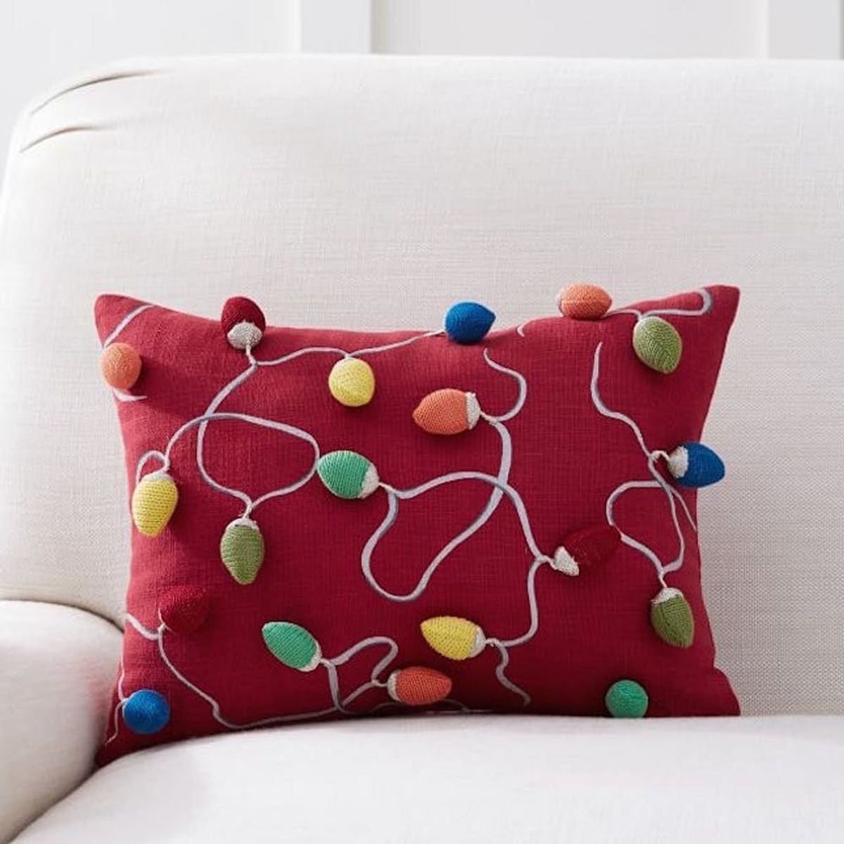 Red pillow with colorful, embroidered Christmas lights design on a white couch.