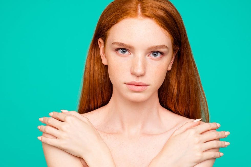 Redhead woman covering her chest in front of a turquoise blue background.