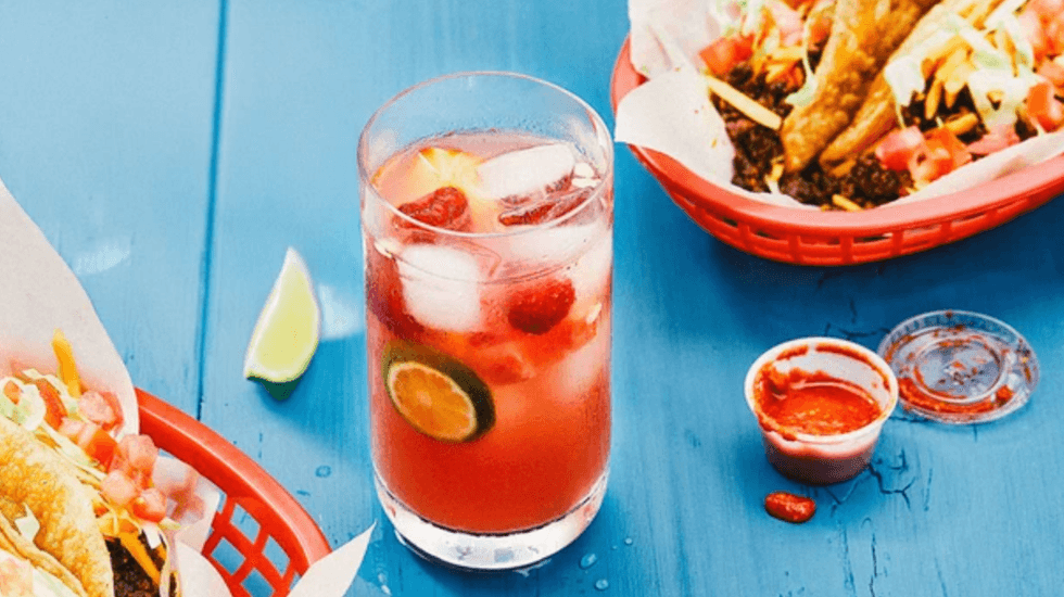 Refreshing pink drink with ice and limes, tacos, and sauce on a blue table.