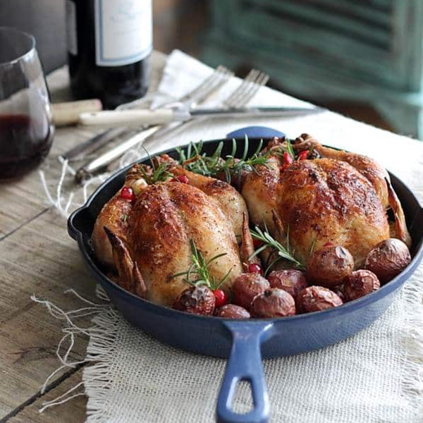 Roasted chickens with potatoes and herbs in a skillet, served with red wine on a rustic table.