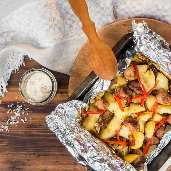 Roasted meat and vegetables in foil, wooden spoon, salt jar, cloth on wooden table.
