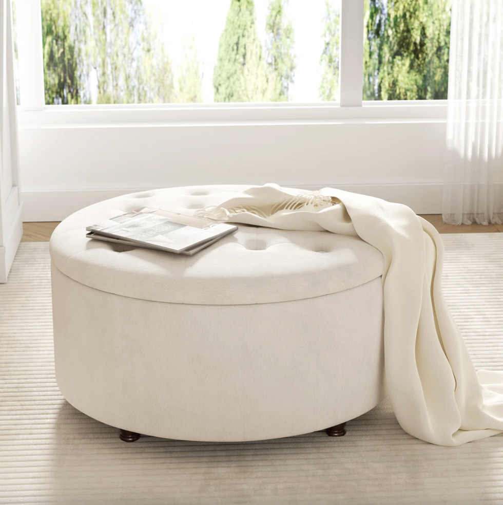 Round beige ottoman with a blanket and magazine, near a window with green trees outside.