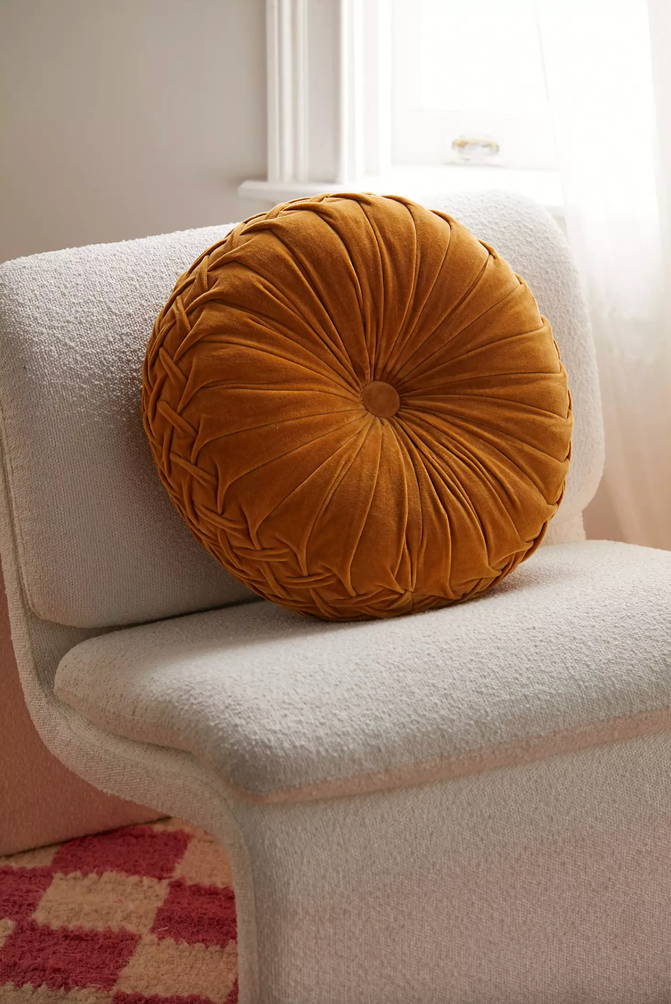 Round mustard pillow on a textured white chair by a window with soft lighting.