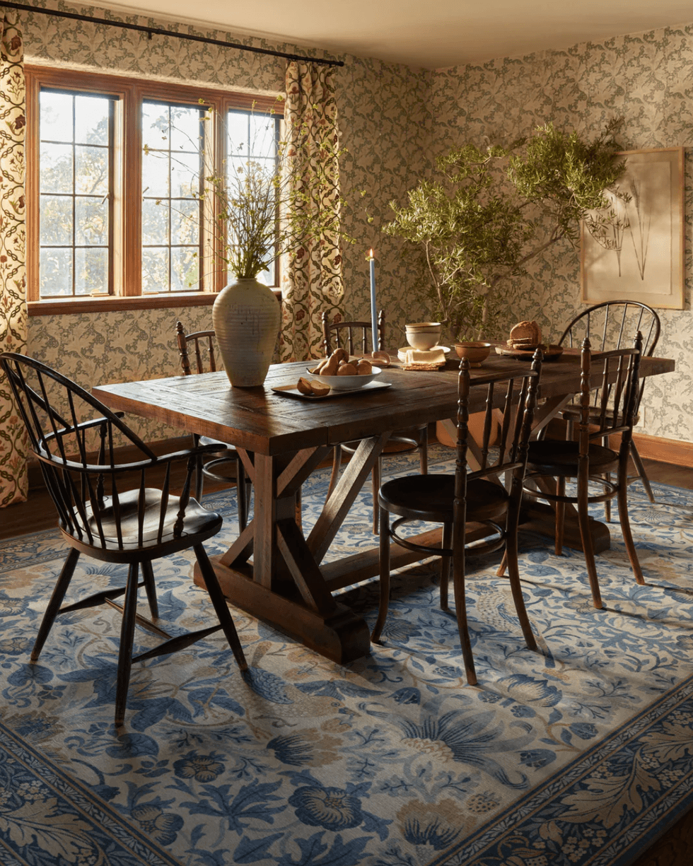 Rustic dining room with a wooden table, chairs, plants, and floral wallpaper.