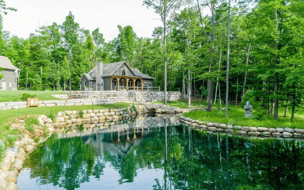 Rustic house by a stone-lined pond, surrounded by lush green trees.