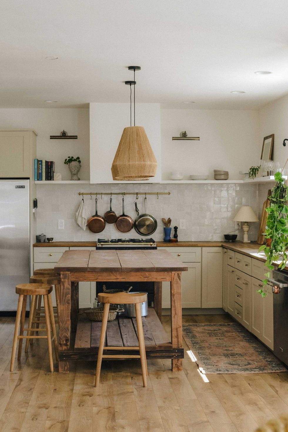 Rustic kitchen with wooden island, hanging pots, wicker pendant, and plants.